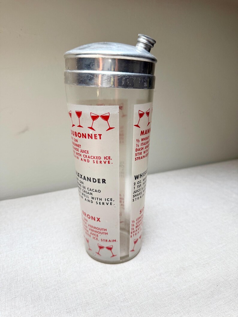 May include: A vintage cocktail shaker with a stainless steel top and a clear glass body. The glass features printed recipes for classic cocktails like Dubonnet and Alexander, with red graphic illustrations.