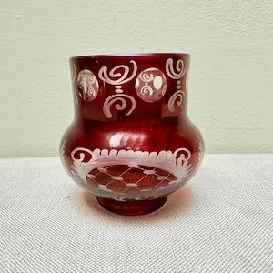May include: A deep red glass vase featuring intricate white etched patterns. The upper section displays a series of circular motifs with swirling lines, while the lower body is decorated with a detailed lattice design and ornate floral-like accents.