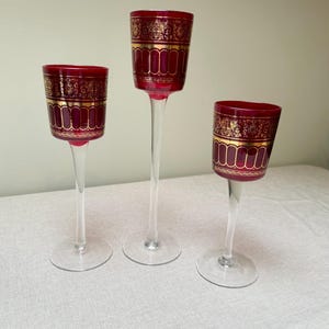Puede incluir: Tres copas de vino con cuencos de color rojo rubí y un diseño dorado. Las copas tienen un tallo transparente y delgado y una base redonda. Las copas son de diferentes alturas. El diseño es ornamentado.