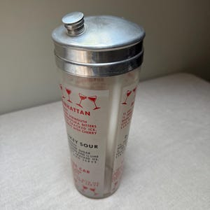 May include: Vintage cocktail shaker with a silver lid and a clear glass body. The glass features red illustrations of cocktail glasses and drink recipes, including "Manhattan" and "Whiskey Sour". A classic barware item.