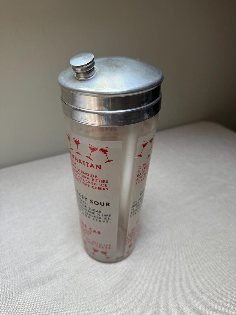 May include: Vintage cocktail shaker with a silver lid and a clear glass body. The glass features red illustrations of cocktail glasses and drink recipes, including "Manhattan" and "Whiskey Sour". A classic barware item.