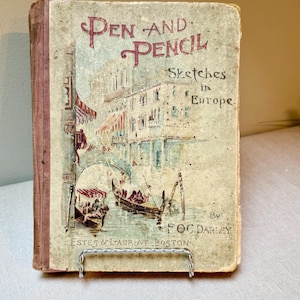 以下が含まれることがあります： FOC Darleyによる「Pen and Pencil Sketches in Europe」というタイトルのアンティーク本。表紙には、ゴンドラのあるヨーロッパの都市の水彩画イラストが描かれています。本は銀色の金属スタンドに展示されています。