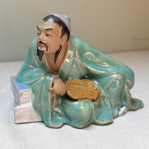 Op de afbeelding: Een keramieken beeldje van een zittende Aziatische man in een turquoise gewaad, met een waaier. De figuur heeft een zwarte baard en zit op een blauw-witte basis. Decoratief item.