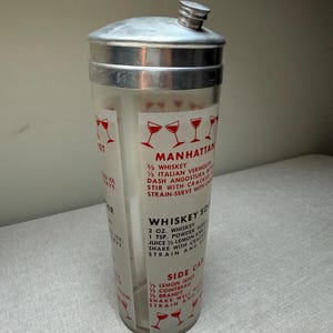 May include: Vintage cocktail shaker with a silver lid and a clear glass body. The shaker features red text and illustrations of cocktail glasses, with recipes for Manhattan, Whiskey Sour, and Side Car cocktails.