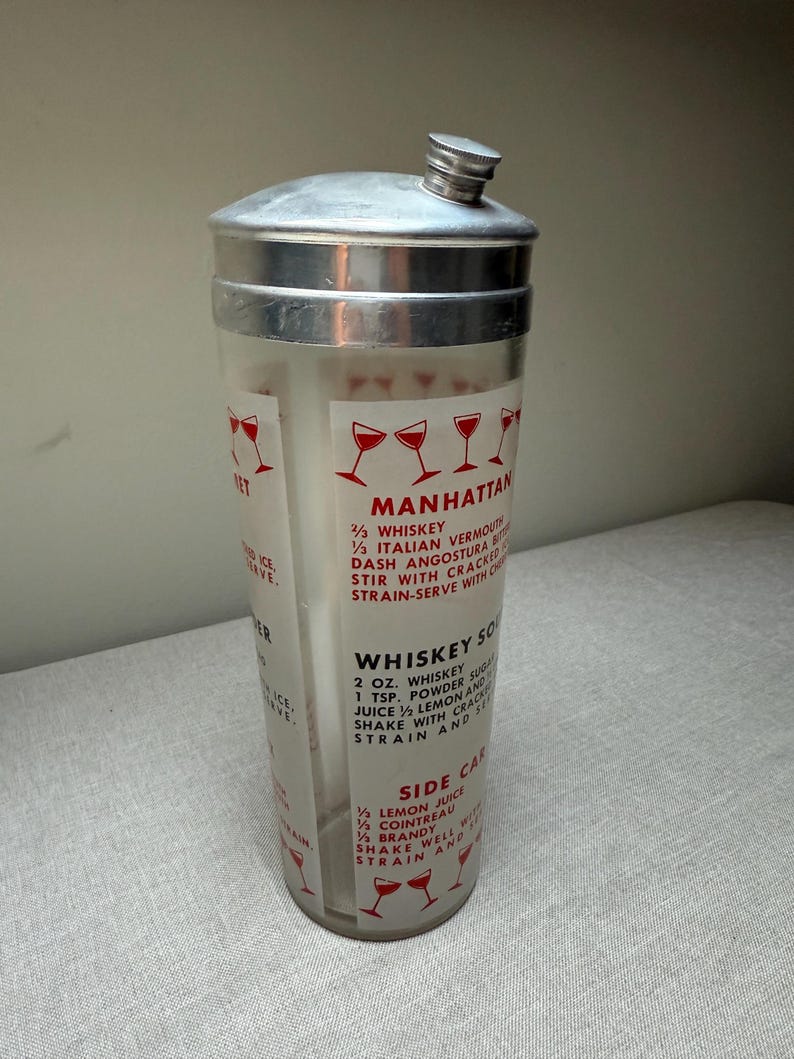 May include: Vintage cocktail shaker with a silver lid and a clear glass body. The shaker features red text and illustrations of cocktail glasses, with recipes for Manhattan, Whiskey Sour, and Side Car cocktails.