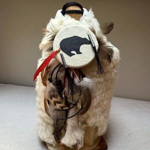Könnte beinhalten: Handgefertigte Puppe mit cremefarbenem, strukturiertem Äußeren, verziert mit Federn und einem kreisförmigen Emblem mit einer schwarzen Bärensilhouette. Die Puppe hat ein schwarzes Stirnband und ist auf einem Holzsockel montiert.