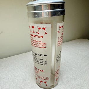 May include: A vintage cocktail shaker with a silver lid and a clear glass body. The glass features red text and illustrations of cocktail glasses, with recipes for Manhattan, Whiskey Sour, and Side Car cocktails.