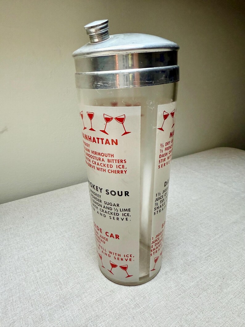 May include: A vintage cocktail shaker with a silver lid and a clear glass body. The glass features red text and illustrations of cocktail glasses, with recipes for Manhattan, Whiskey Sour, and Side Car cocktails.