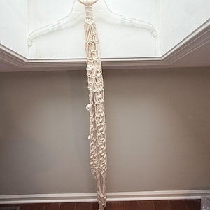 Puede incluir: Un colgador de plantas de macramé color crema con un anillo de madera. El diseño largo y tejido presenta intrincados nudos y una borla en la parte inferior. Esta pieza decorativa está diseñada para sostener una planta en maceta.