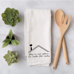 May include: White kitchen towel with a black house graphic and the text "This is our place, we make the rules." Two wooden cooking utensils are crossed on the right. Green succulents in small pots are arranged around the towel.