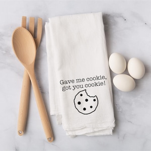 May include: A white kitchen towel with the text "Gave me cookie, got you cookie!" and a cookie graphic. Two wooden spoons and three white eggs are also in the image. The items are arranged on a marble surface.