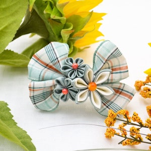 May include: A handcrafted hair clip featuring a plaid bow design in shades of blue, white, and orange. The clip is adorned with fabric flowers in complementary colors, with orange and red accents. The accessory is set against a backdrop of sunflowers and foliage.