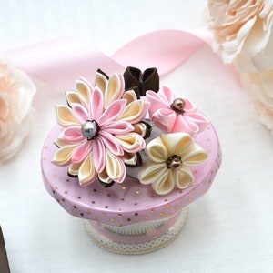 Puede incluir: Una pinza para el pelo floral de color rosa y crema con un centro de perla, expuesta sobre una base de lunares rosa y blanco con detalles dorados.