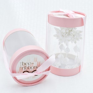 May include: White hair comb with a floral design, packaged in a clear cylinder with a pink lid and a pink ribbon. The cylinder has a label that says "bee-ribbone".