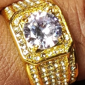 May include: A gold-coloured ring featuring a large, clear, square-cut central stone. The ring is embellished with many small, clear stones around the centre and on the band, producing a sparkling effect. The ring has a striking, rectangular design.