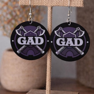 May include: A pair of black circular earrings featuring the word "GAD" in white, and a purple axe and weapon design. The earrings are suspended from silver hooks and displayed on a wooden stand.