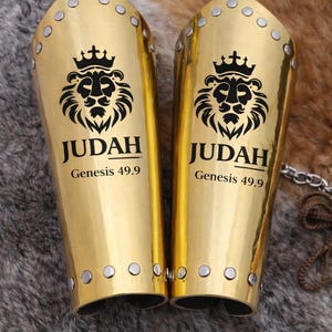 May include: Two gold-colored arm guards with a lion crest and the word "JUDAH" printed in black. The text "Genesis 49.9" is below the word "JUDAH". The arm guards are decorated with silver rivets.