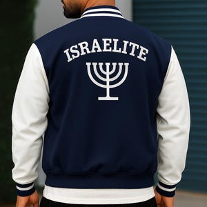 May include: Navy blue and white varsity jacket with the word "ISRAELITE" and a menorah symbol on the back. The jacket has white sleeves and striped cuffs and collar. The person is wearing a white t-shirt and dark blue jeans.