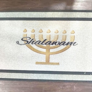 May include: Rectangular doormat with a light beige center, a dark gray border, and a textured appearance. The mat features a gold menorah design and the word "Shalawam" in black script, suitable for a home entrance.