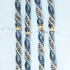 May include: Four macrame keyrings with bronze-coloured clasps. Each keyring features a spiral design in blue, beige, and white, with a gold-coloured crown accent. The tassels at the bottom are blue, beige, and white.