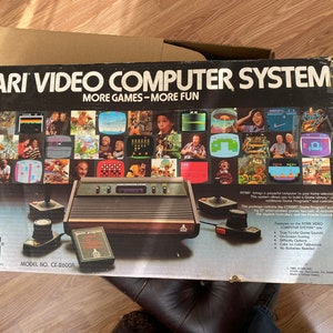 May include: An Atari Video Computer System box with the text "ATARI VIDEO COMPUTER SYSTEM MORE GAMES - MORE FUN." The box features images of various games and includes a console, two joysticks, and a game cartridge.