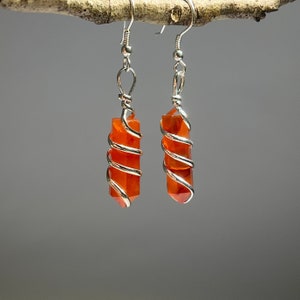 May include: A pair of silver wire wrapped carnelian earrings. The carnelian stones are a deep orange color and are wrapped in silver wire that spirals around the stones.