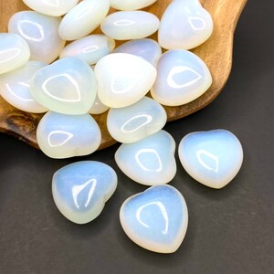 A group of white heart-shaped opalite stones. The stones are smooth and polished, with a slight iridescent sheen. They are arranged in a pile on a wooden surface.
