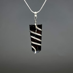 May include: A black crystal pendant wrapped in silver-colored wire, suspended from a silver chain. The crystal is rectangular with a pointed end. The wire is wrapped around the crystal in a spiral pattern.