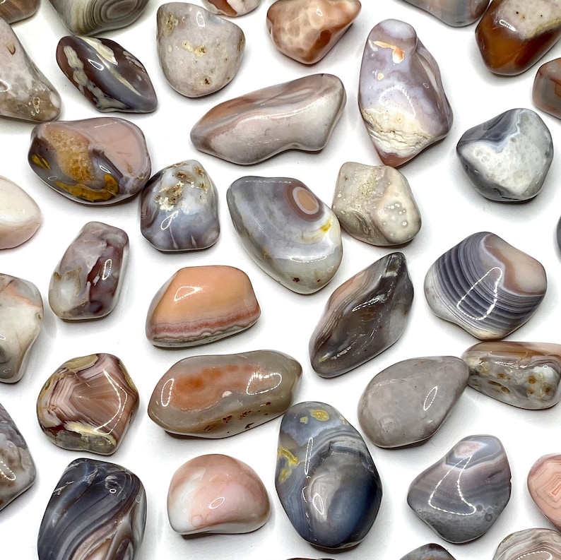A collection of tumbled agate stones in various shades of pink, gray, and white. The stones are smooth and polished, with intricate patterns of banding and swirls.
