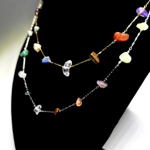 May include: A layered necklace with a silver chain and a gold chain. The necklace features a variety of colourful gemstones, including clear, blue, green, pink, red, and purple.