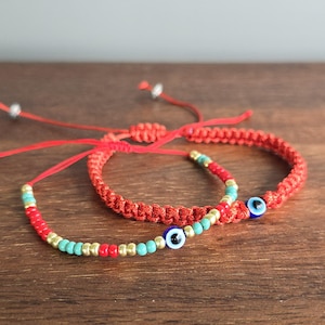 May include: Two adjustable bracelets featuring red cord and colorful beads. One bracelet has a braided red cord, while the other is a thin string with alternating gold, turquoise, and red beads. Both bracelets include a blue, white, and black eye bead.