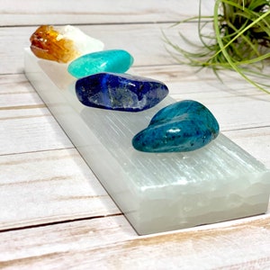 A selenite charging bar with five tumbled stones. The stones are orange, white, teal, blue, and green. The selenite plate is white and has a striated texture.