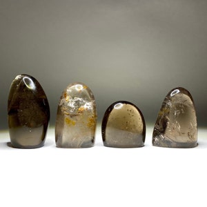 May include: Four smooth, polished smoky quartz stones, each with a unique shape and a slightly different shade of brown. The stones are arranged in a row on a white surface.