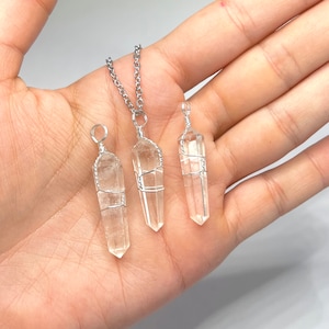 May include: Three clear quartz crystal pendants wrapped in silver wire. The pendants are hanging from silver chains. The pendants are different sizes and are displayed on a hand.