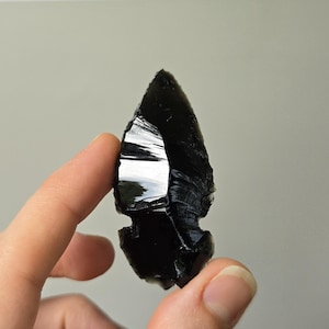 May include: A black obsidian arrowhead featuring a sharp, pointed tip and a notched base. The surface of the stone is smooth and reflective, displaying distinct conchoidal fracture patterns along the edges, characteristic of knapped volcanic glass.