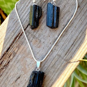 Puede incluir: Un conjunto de collar y pendientes de turmalina negra. El collar presenta un largo colgante de cristal negro en bruto en una cadena de plata. Los pendientes tambi&eacute;n est&aacute;n hechos con cristales de turmalina negra en bruto.