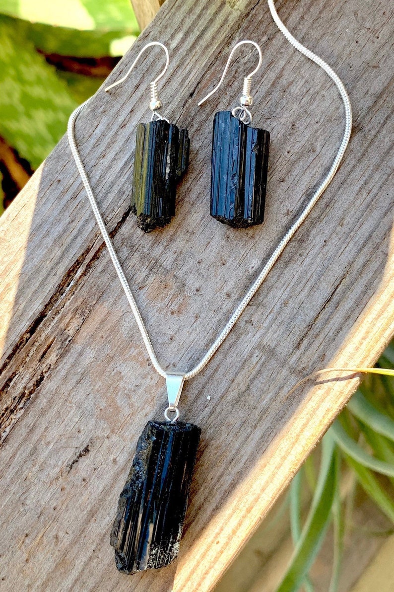 Puede incluir: Un conjunto de collar y pendientes de turmalina negra. El collar presenta un largo colgante de cristal negro en bruto en una cadena de plata. Los pendientes tambi&eacute;n est&aacute;n hechos con cristales de turmalina negra en bruto.