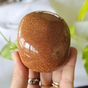 Könnte beinhalten: Ein polierter, oval geformter goldener Sandstein-Edelstein. Der Stein hat ein funkelndes, glitzerndes Aussehen und wird in einer Hand gehalten. Der Edelstein hat einen warmen, erdigen Farbton und reflektiert das Licht. Der Hintergrund ist verschwommen, mit grünem Laub sichtbar.