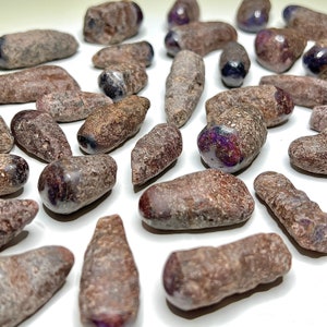 Peut inclure: Une collection de pierres de rubis brutes, de couleur violette. Les pierres sont de tailles et de formes variées, certaines ayant une pointe pointue. Les pierres sont présentées sur une surface blanche.