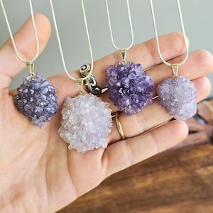 May include: Four amethyst crystal pendants on silver chains. The crystals vary in shades of purple and lavender. Each pendant is a cluster of small, sparkling crystals. The necklaces are displayed on a hand.