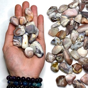 A collection of tumbled agate stones in shades of pink, brown, white, and gray. The stones are smooth and polished, with intricate patterns of banding and swirls.