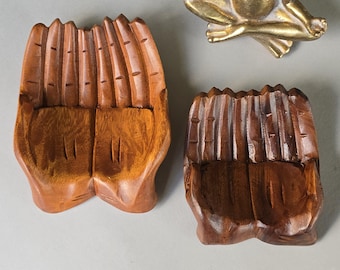 May include: Two carved wooden bowls shaped like cupped hands, one in a light brown finish and one in a dark brown finish, accompanied by a small gold-colored metallic frog figurine in a seated meditation pose with its legs crossed and hands resting.