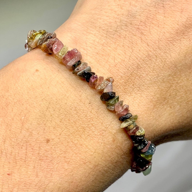 A bracelet made of small, multi-colored, chipped gemstones. The gemstones are in shades of pink, green, black, and brown. The bracelet is strung on a clear elastic cord.