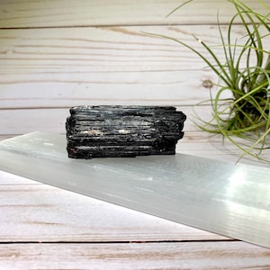 A black tourmaline crystal on a selenite charging bar. The crystal is a dark gray color with a rough, natural texture. The selenite plate is a smooth, white color.