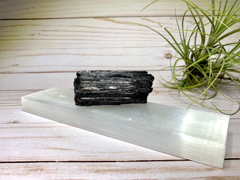 A black tourmaline crystal on a selenite charging bar. The crystal is a dark gray color with a rough, natural texture. The selenite plate is a smooth, white color.