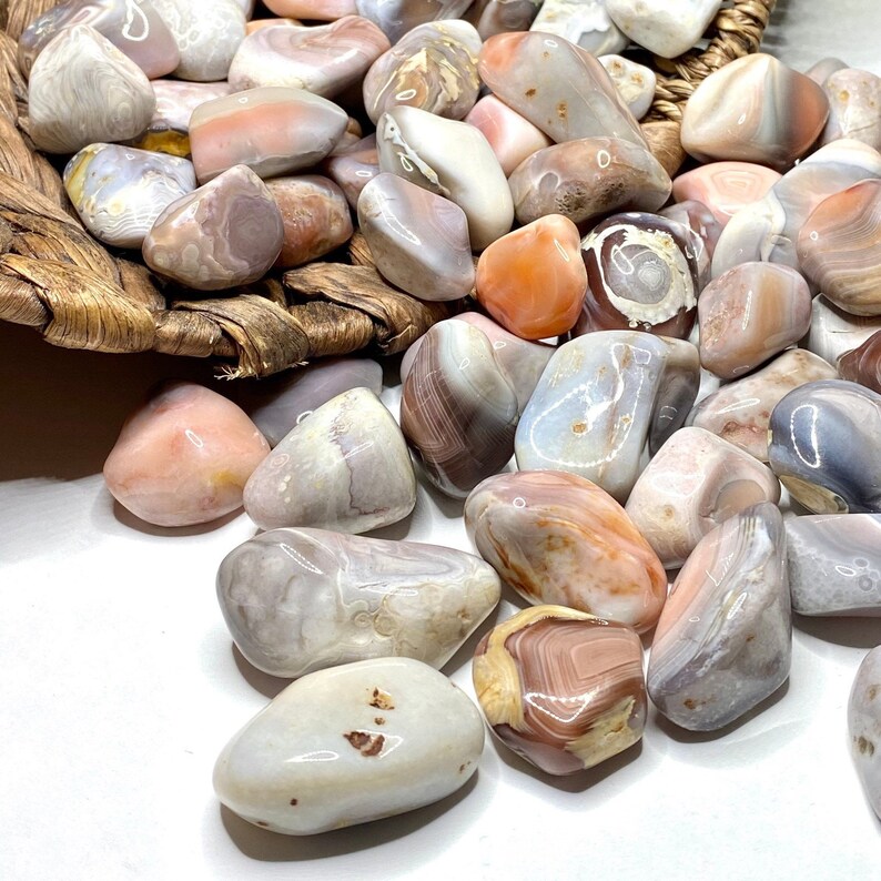 A collection of tumbled agate stones in various shades of pink, white, and brown. The stones are smooth and polished, with intricate patterns of banding and swirls. The stones are arranged on a white surface.