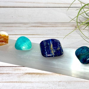 A selenite charging plate with four tumbled stones: a brown calcite, a green amazonite, a blue lapis lazuli, and a blue apatite.