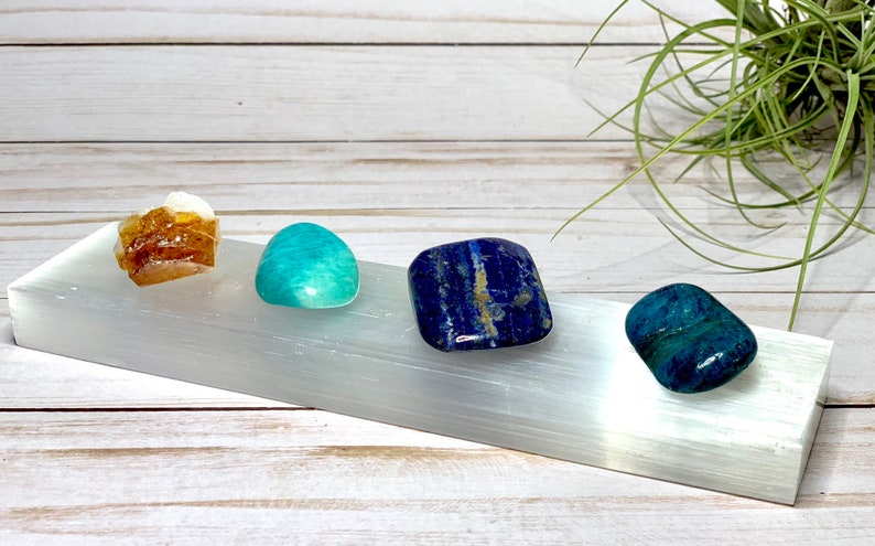 A selenite charging plate with four tumbled stones: a brown calcite, a green amazonite, a blue lapis lazuli, and a blue apatite.