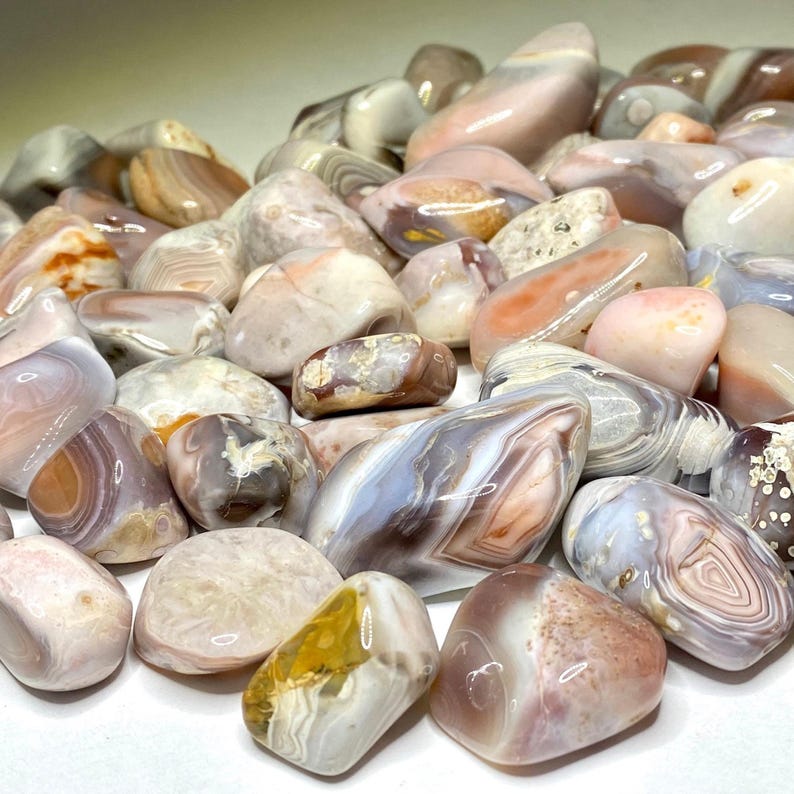 A collection of tumbled agate stones in various shades of pink, white, and brown. The stones have a banded pattern and are smooth and polished.