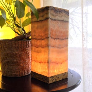 May include: A rectangular table lamp crafted from translucent banded stone with horizontal layers of orange, cream, and dark brown. The lamp features a solid, textured stone base and emits a warm, soft glow from within its polished, natural stone structure.